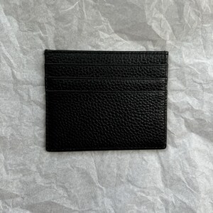 May include: A black leather card holder with multiple card slots. The card holder has a textured surface and is rectangular in shape. The card holder is displayed on a white, textured background.