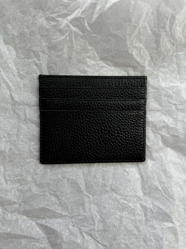 May include: A black leather card holder with multiple card slots. The card holder has a textured surface and is rectangular in shape. The card holder is displayed on a white, textured background.
