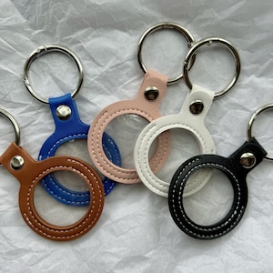 May include: A collection of keychains in various colors, including brown, blue, pink, white, and black. Each keychain features a circular leather holder with a stitched border and a silver metal ring for attaching keys.