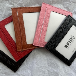 May include: A collection of leather cardholders in red, brown, pink, and black. Each cardholder has a clear window and a slot for cards. The black cardholder displays a white card with "RFID PROTECTED" text.