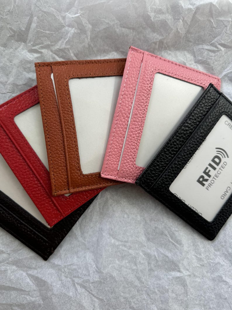 May include: A collection of leather cardholders in red, brown, pink, and black. Each cardholder has a clear window and a slot for cards. The black cardholder displays a white card with "RFID PROTECTED" text.