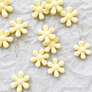 May include: A collection of pale yellow, six-petaled flower charms, each featuring a smooth, glossy finish and a thin, flexible wire loop extending from the base, arranged in a scattered pattern across the frame for decorative use.