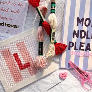 May include: A craft scene with embroidery floss in red, pink, and cream, along with a needle, scissors, and a canvas with the letter "L" stitched in red. A notepad with the text "MO NDLE PLEA" is also visible.