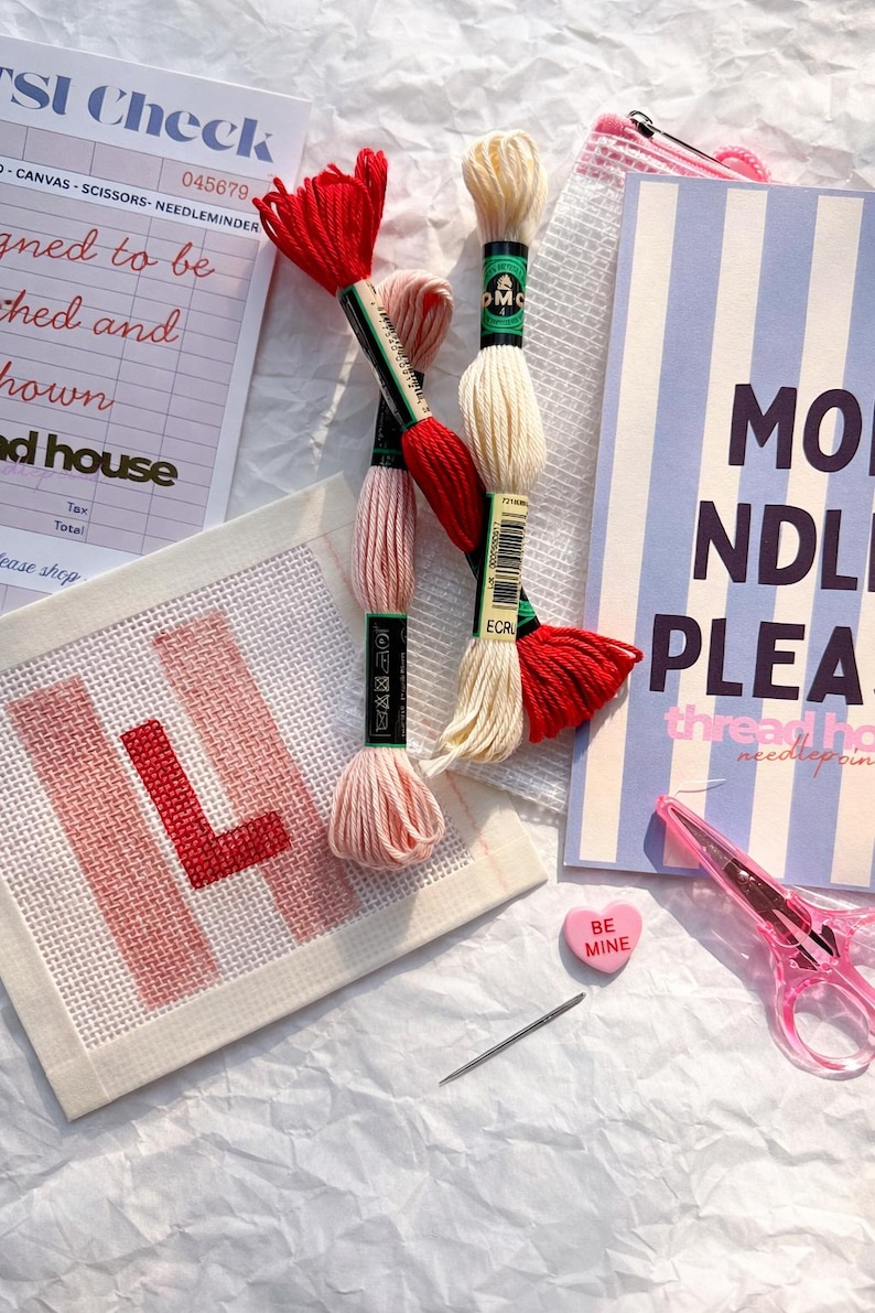 Puede incluir: Una escena de manualidades con hilo de bordar rojo, rosa y crema, una aguja, tijeras y un lienzo con la letra "L" bordada en rojo. Tambi&eacute;n se ve un bloc de notas con el texto "MO NDLE PLEA".