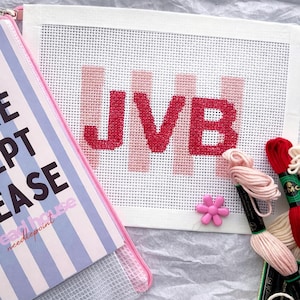 May include: A needlepoint canvas with the letters "JVB" in red, with pink vertical stripes. A pink flower, embroidery floss, and a striped case with text are also visible. The case has a pink zipper.