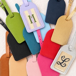 May include: A collection of luggage tags in various colors including lime green, lavender, navy blue, tan, black, red, pink, and silver. Two tags feature needlepoint inserts with the letters M and L stitched in dark green and black thread.