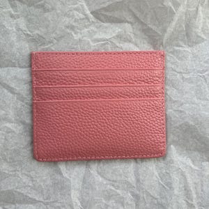 May include: A pink leather card holder with multiple card slots. The card holder is rectangular and has a textured surface. The stitching is visible around the edges.