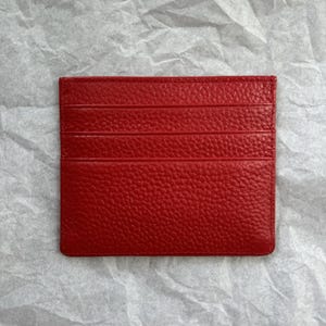 May include: A bright red leather card holder with multiple card slots. The card holder has a textured surface and is rectangular in shape. The card holder is displayed on a white, textured background.