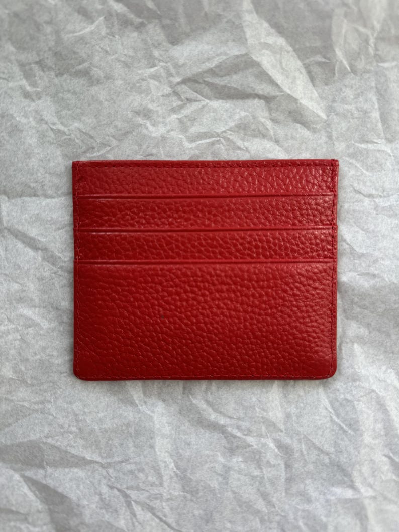May include: A bright red leather card holder with multiple card slots. The card holder has a textured surface and is rectangular in shape. The card holder is displayed on a white, textured background.