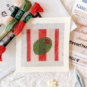 May include: A square needlepoint canvas featuring a green olive with a red pimento center against a background of vertical red stripes. The kit includes three skeins of embroidery floss in pink, green, and red, a needle, and a small pair of scissors.