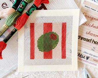 May include: A square needlepoint canvas featuring a green olive with a red pimento center against a background of vertical red stripes. The kit includes three skeins of embroidery floss in pink, green, and red, a needle, and a small pair of scissors.