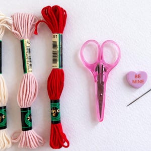 Puede incluir: Un conjunto de suministros de bordado, que incluye tres madejas de hilo en crema, rosa y rojo, unas tijeras rosas, una aguja y un caramelo en forma de coraz&oacute;n con el texto "BE MINE".