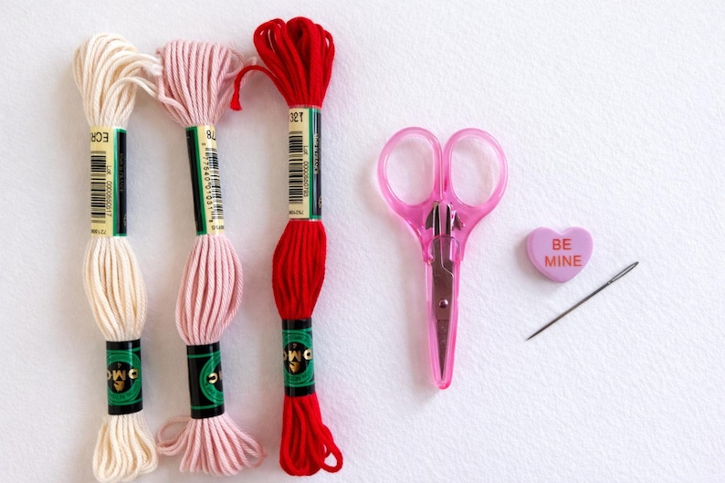 Puede incluir: Un conjunto de suministros de bordado, que incluye tres madejas de hilo en crema, rosa y rojo, unas tijeras rosas, una aguja y un caramelo en forma de coraz&oacute;n con el texto "BE MINE".