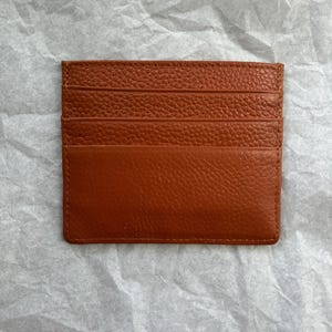 May include: A rectangular, brown leather card holder with multiple card slots. The card holder is made of textured leather and has a stitched border. The card holder is designed to hold credit cards, business cards, or other small items.