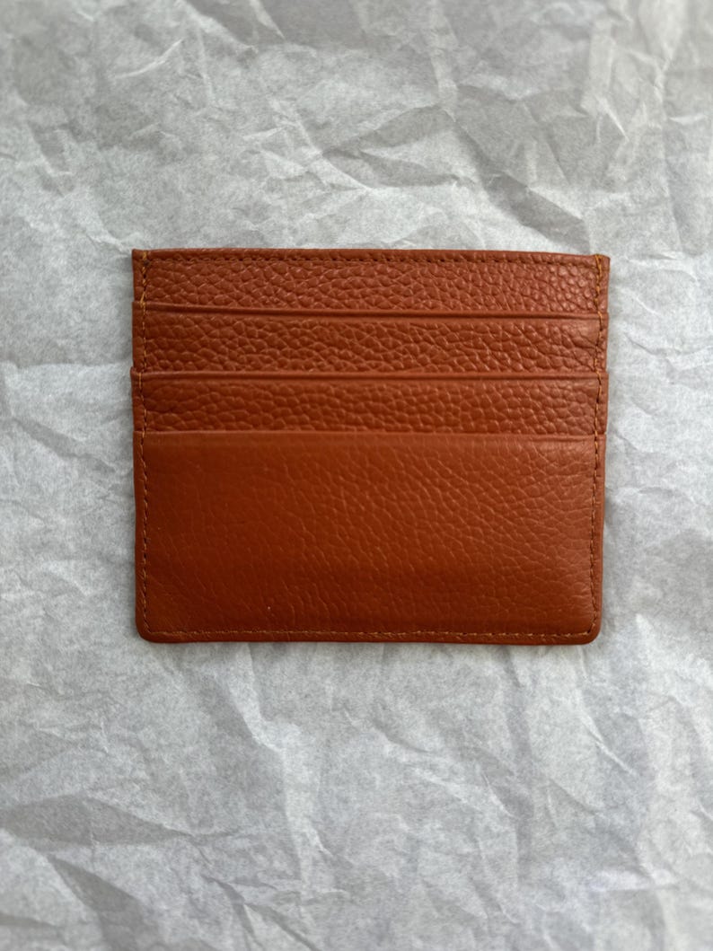 May include: A rectangular, brown leather card holder with multiple card slots. The card holder is made of textured leather and has a stitched border. The card holder is designed to hold credit cards, business cards, or other small items.