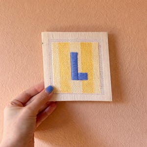 Puede incluir: Un bordado de punto de aguja cuadrado hecho a mano con la letra "L" en azul, sobre un fondo de rayas verticales amarillas y blancas. El bordado se muestra contra una pared de color melocot&oacute;n.