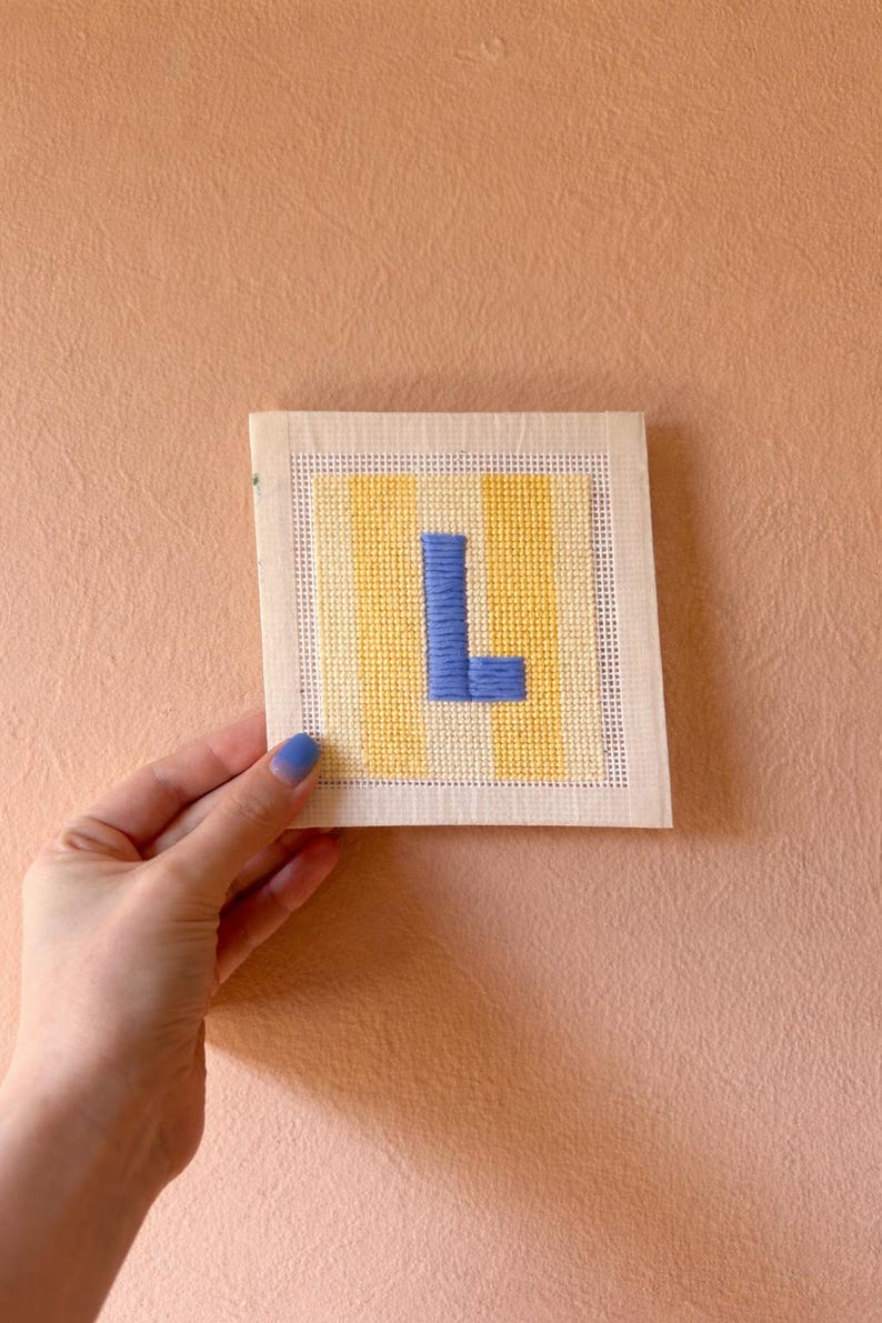 Puede incluir: Un bordado de punto de aguja cuadrado hecho a mano con la letra "L" en azul, sobre un fondo de rayas verticales amarillas y blancas. El bordado se muestra contra una pared de color melocot&oacute;n.
