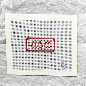 May include: A rectangular white needlepoint canvas featuring the red stitched text USA centered within a red rectangular border. A small blue stitched monogram logo is located in the bottom right corner of the grid, which is composed of evenly spaced holes for embroidery work.