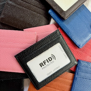 May include: A collection of leather cardholders in various colors, including black, pink, brown, blue, and orange. One black cardholder displays a credit card with "RFID PROTECTED" text. The cardholders have multiple card slots and a clear window.