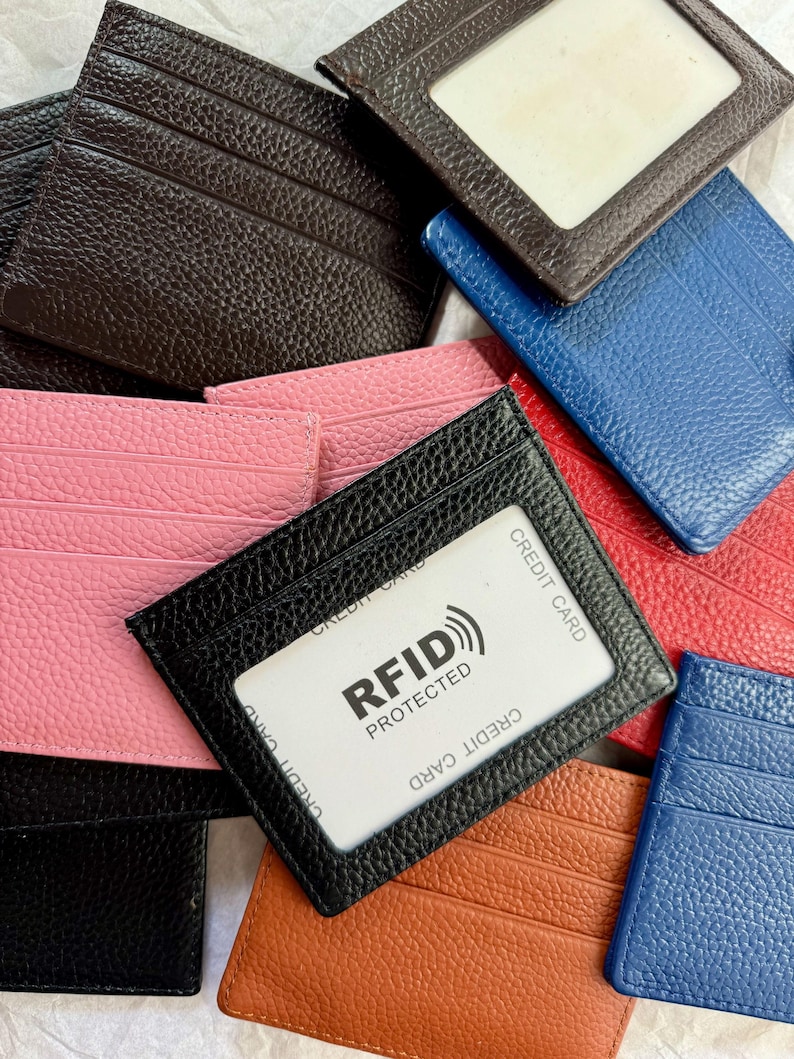 May include: A collection of leather cardholders in various colors, including black, pink, brown, blue, and orange. One black cardholder displays a credit card with "RFID PROTECTED" text. The cardholders have multiple card slots and a clear window.