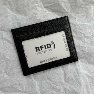May include: Black leather card holder with a clear window displaying a white card. The white card has the text "RFID PROTECTED" and "CREDIT CARD" printed on it. The card holder is rectangular and has a textured surface.