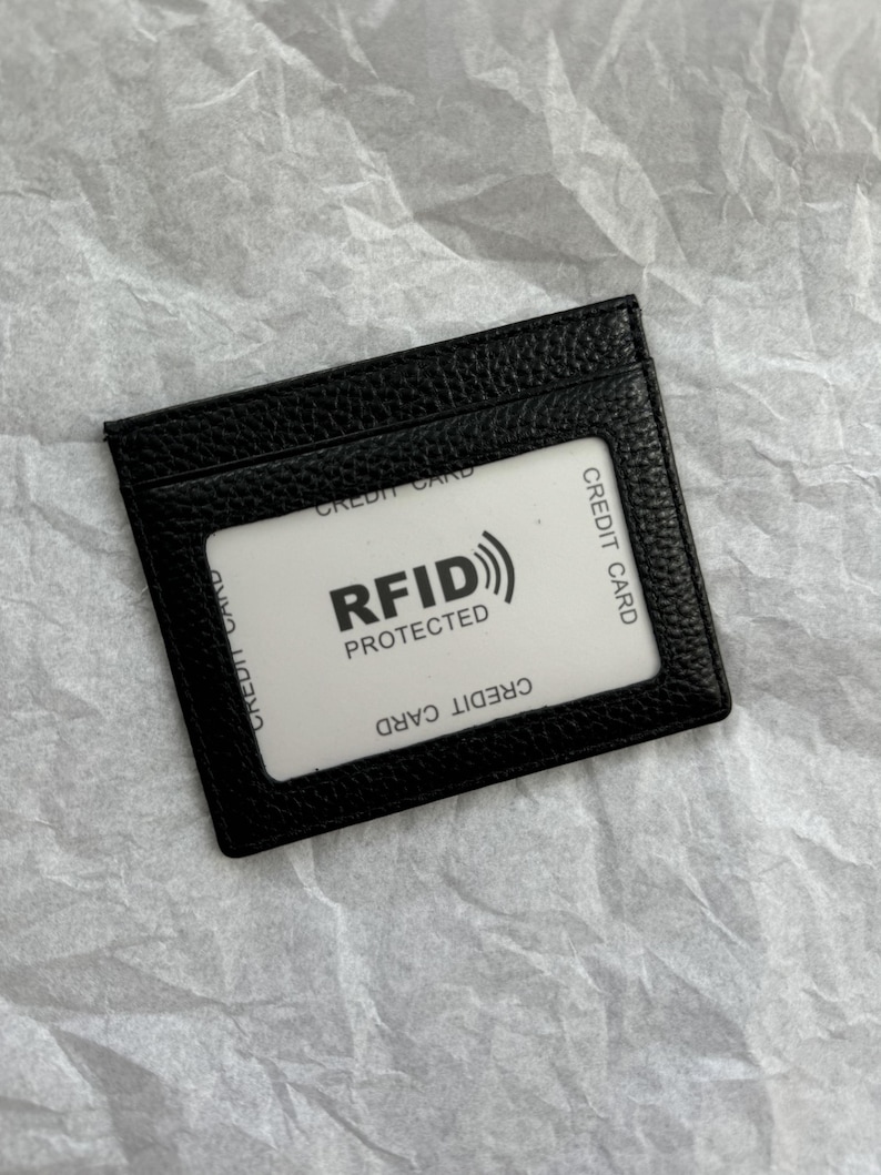 May include: Black leather card holder with a clear window displaying a white card. The white card has the text "RFID PROTECTED" and "CREDIT CARD" printed on it. The card holder is rectangular and has a textured surface.