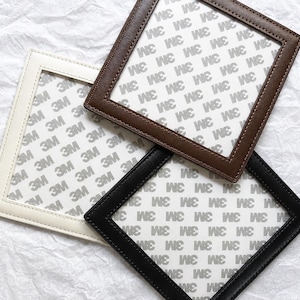 May include: Three square frames with stitched borders in white, brown, and black. Each frame features a white adhesive backing paper printed with a repeating 3M logo pattern. The frames are arranged in an overlapping, layered composition.