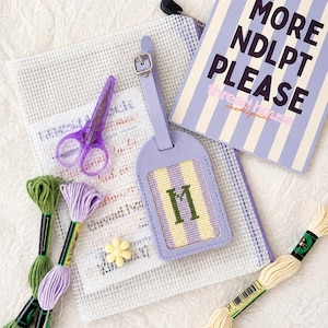 May include: A lavender luggage tag with a needlepoint insert featuring the letter M, a purple pair of scissors, a white mesh pouch, a yellow flower-shaped bead, three skeins of embroidery floss, and a book titled MORE NDLPT PLEASE.