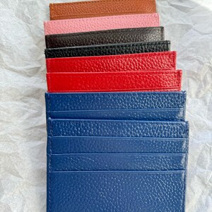 May include: A stack of colorful leather cardholders in various colors, including brown, pink, black, red, and blue. Each cardholder has multiple slots for cards and a textured surface. The cardholders are arranged on a white, crumpled paper background.
