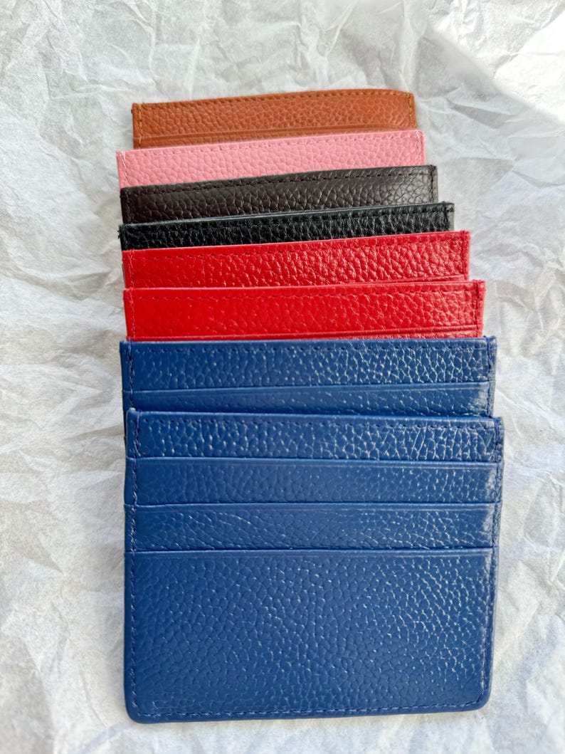 May include: A stack of colorful leather cardholders in various colors, including brown, pink, black, red, and blue. Each cardholder has multiple slots for cards and a textured surface. The cardholders are arranged on a white, crumpled paper background.