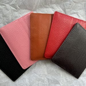 May include: A collection of five leather cardholders in various colors: black, pink, brown, red, and dark brown. Each cardholder has a textured surface and a rectangular shape, suitable for carrying credit cards or IDs.