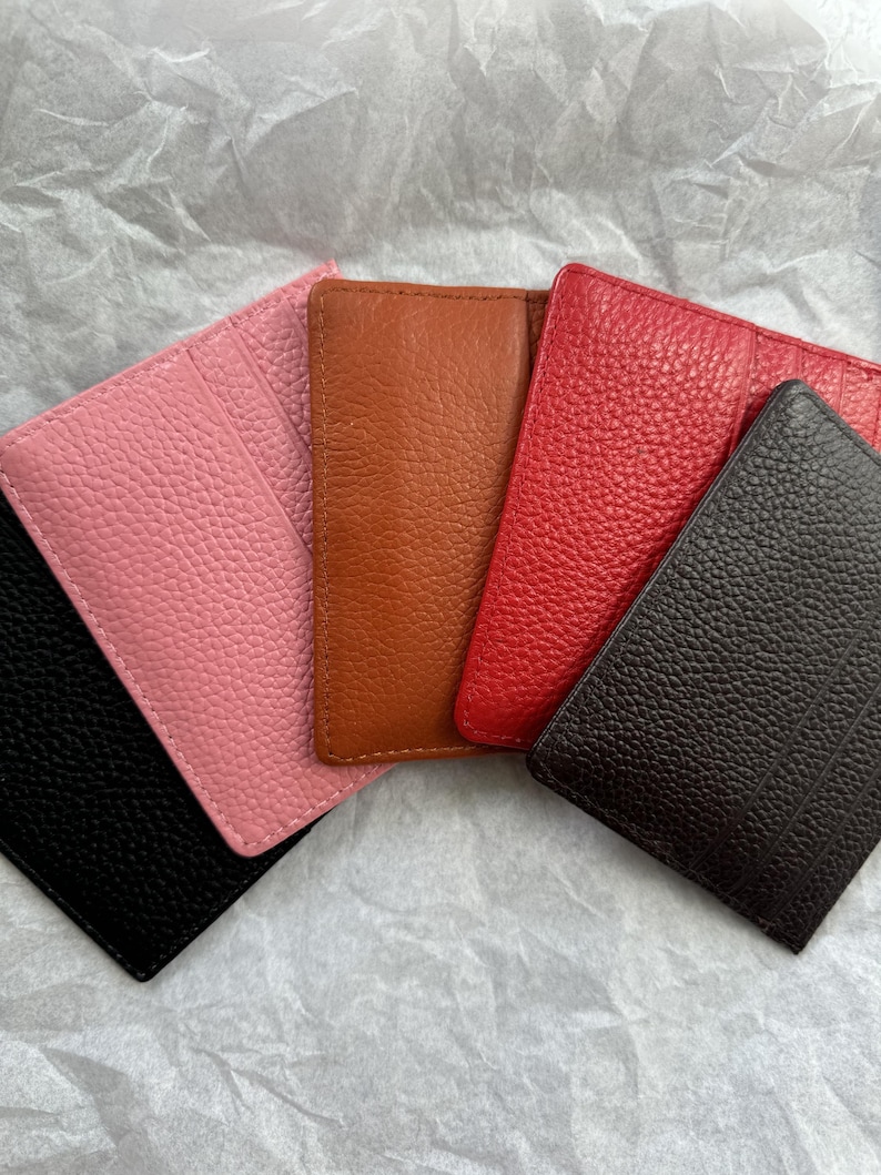 May include: A collection of five leather cardholders in various colors: black, pink, brown, red, and dark brown. Each cardholder has a textured surface and a rectangular shape, suitable for carrying credit cards or IDs.