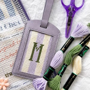 May include: A lavender luggage tag with a stitched "M" on a striped background, next to purple scissors and spools of embroidery floss in various colors. A clear mesh bag and a small flower are also visible.