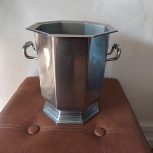 Peut inclure: Seau à glace octogonal argenté avec deux poignées incurvées. Le seau a un design géométrique et repose sur un pouf en cuir marron. Le seau est en métal et a une finition brillante.