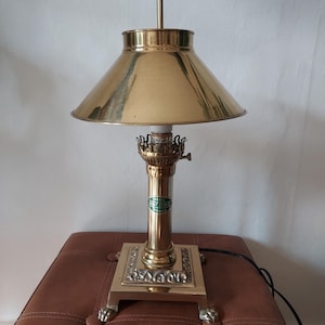 Puede incluir: Una lámpara de escritorio de latón con una pantalla cónica y una base cuadrada con detalles decorativos. La lámpara tiene un cuerpo cilíndrico y una pequeña etiqueta verde. La lámpara está sobre un otomán de cuero marrón.