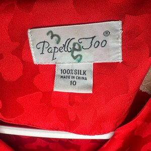 Peut inclure: Gros plan sur un v&ecirc;tement en soie rouge vif avec un motif floral. L'&eacute;tiquette indique "Papell & Coo" et "100% Soie, Fabriqu&eacute; en Chine, 10". Le tissu est d'un rouge riche et uni.