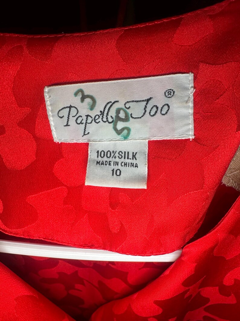 Peut inclure: Gros plan sur un v&ecirc;tement en soie rouge vif avec un motif floral. L'&eacute;tiquette indique "Papell & Coo" et "100% Soie, Fabriqu&eacute; en Chine, 10". Le tissu est d'un rouge riche et uni.