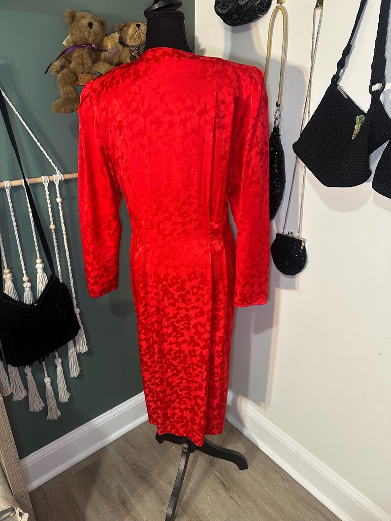 Peut inclure: Une robe rouge vif avec un motif en forme de c&oelig;ur. La robe &agrave; manches longues est pr&eacute;sent&eacute;e sur un mannequin noir. La robe a un ourlet jusqu'aux genoux et une silhouette ajust&eacute;e. Le tissu semble &ecirc;tre une mati&egrave;re soyeuse.