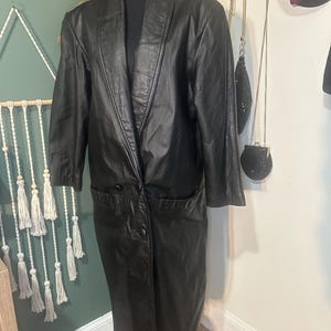 Puede incluir: Un abrigo largo de cuero negro con solapas y dos bolsillos delanteros. El abrigo está abotonado y tiene un estilo clásico y vintage. El abrigo se exhibe en un maniquí.