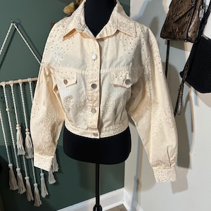 May include: Cream-colored cropped jacket with a button-up front, two chest pockets, and long sleeves. The jacket features a delicate floral pattern and small silver studs. The jacket is displayed on a black mannequin.