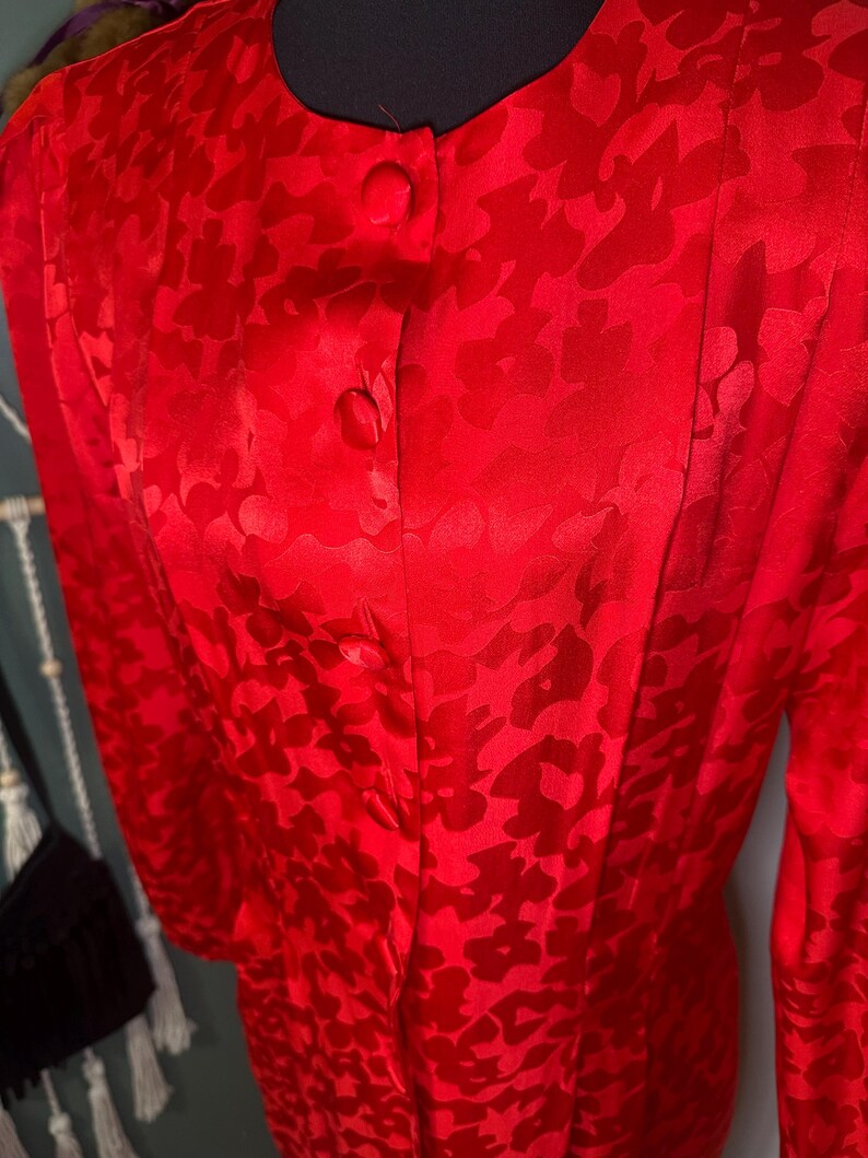 Peut inclure: Une blouse rouge vif &agrave; motifs floraux. Ce haut &agrave; manches longues pr&eacute;sente un col rond et trois boutons visibles. Le tissu a un l&eacute;ger &eacute;clat et le motif floral est l&eacute;g&egrave;rement plus fonc&eacute; que la couleur de base.