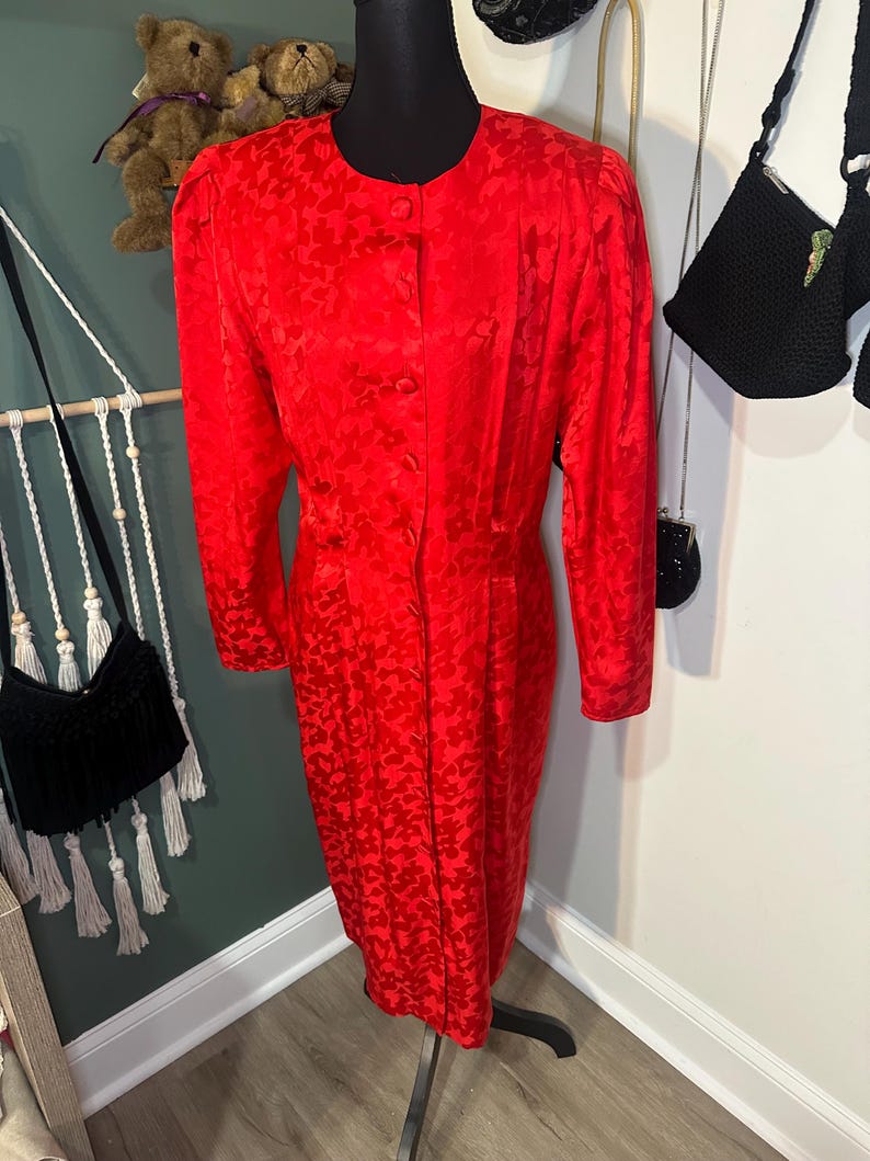 Peut inclure: Une robe rouge vif avec un motif floral subtil. La robe a des manches longues, un devant boutonn&eacute; et un ourlet jusqu'aux genoux. La robe est pr&eacute;sent&eacute;e sur un mannequin noir.