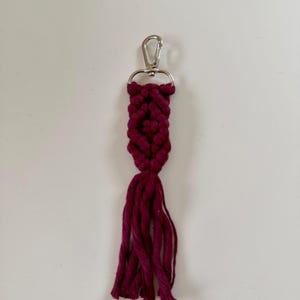 Peut inclure: Un porte-clés macramé fait à la main de couleur bordeaux foncé. Il présente un motif tissé et un fermoir argenté. Une touffe de fils de la même couleur bordeaux orne le bas du porte-clés.