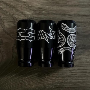 May include: Three black metal objects with white etched designs. The designs include a chain, barbed wire, and a snake. The objects are cylindrical with a flared base and a slightly wider top.