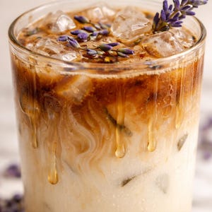 May include: A refreshing iced beverage in a clear glass, featuring layers of creamy white and dark brown liquids, ice cubes, and sprigs of purple lavender. The drink has a drippy, textured appearance, suggesting a sweet, cold treat.
