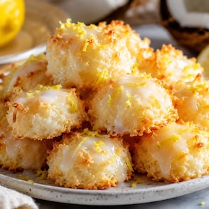 Può includere: Un piatto di macaron al cocco dorati, cosparsi di glassa bianca e scorza di limone. I biscotti sono disposti su un piatto colorato chiaro, con un limone e una noce di cocco sullo sfondo. Un dessert delizioso.