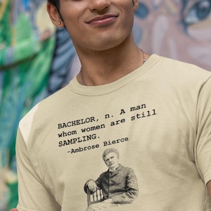 May include: A light beige t-shirt with black text that reads "BACHELOR, n. A man whom women are still SAMPLING." and a small illustration of a man. The quote is attributed to Ambrose Bierce.