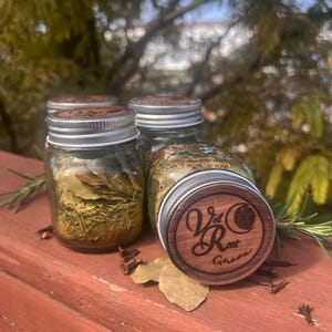 May include: Three clear glass jars filled with dried green herbs and bay leaves. Each jar has a silver metal lid topped with a circular wooden inlay engraved with the text Veil Root Grove and a small pentagram symbol inside a crescent moon.