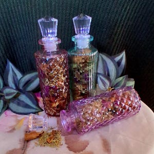 May include: Three decorative glass bottles with crystal stoppers. One is purple, one is amber, and one is green. The bottles are filled with dried herbs and flowers. The purple bottle is lying on its side, with its stopper removed.