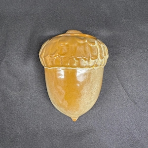 May include: A ceramic acorn-shaped wall pocket with a glossy, honey-brown glaze. The upper cap section features a textured, scalloped design, while the lower nut portion has a smooth, rounded surface. The piece is finished with a small point.